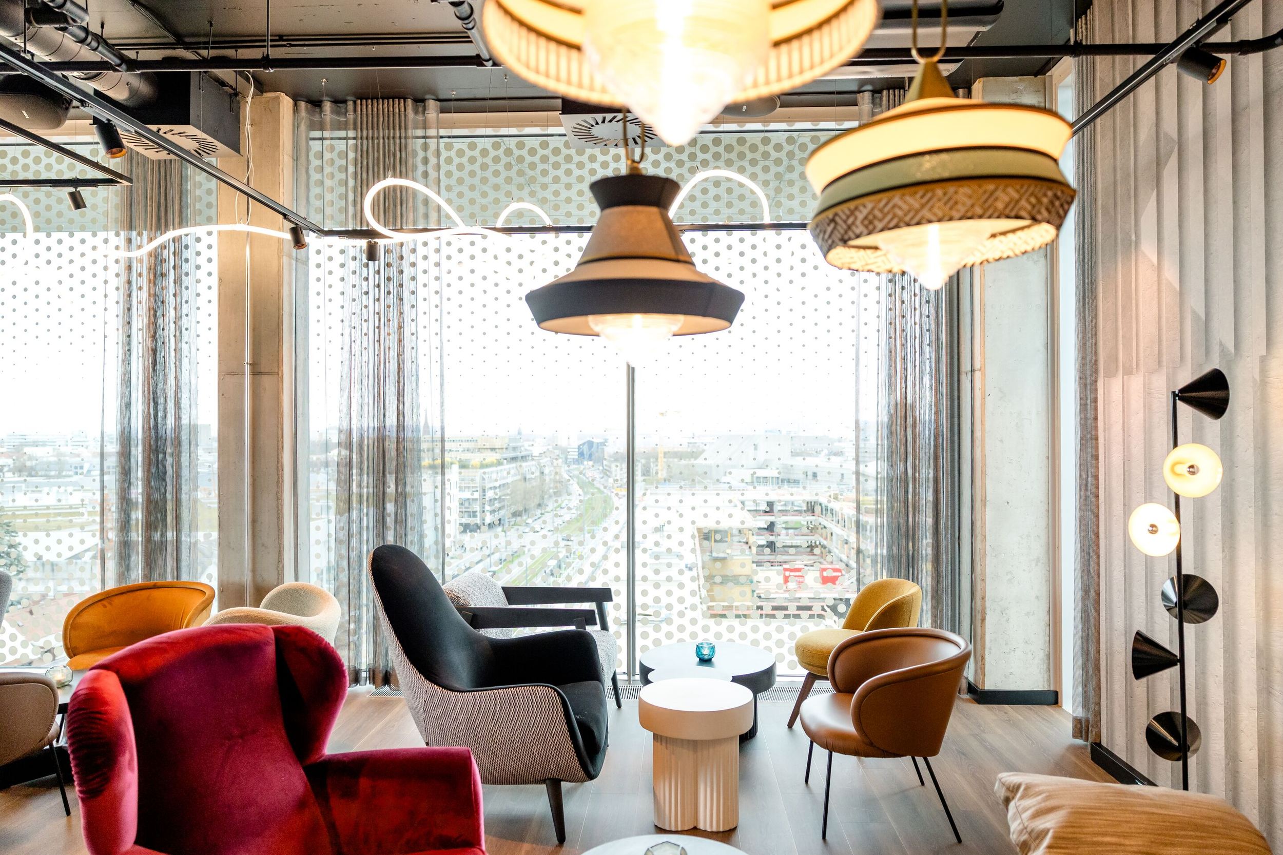 Hotel Karlsruhe Motel One Rooftop Bar mit bequemen Sesseln in warmen Farben, großen Fenstern mit Blick auf die Stadt und stilvollen Hängeleuchten für entspannte Atmosphäre.