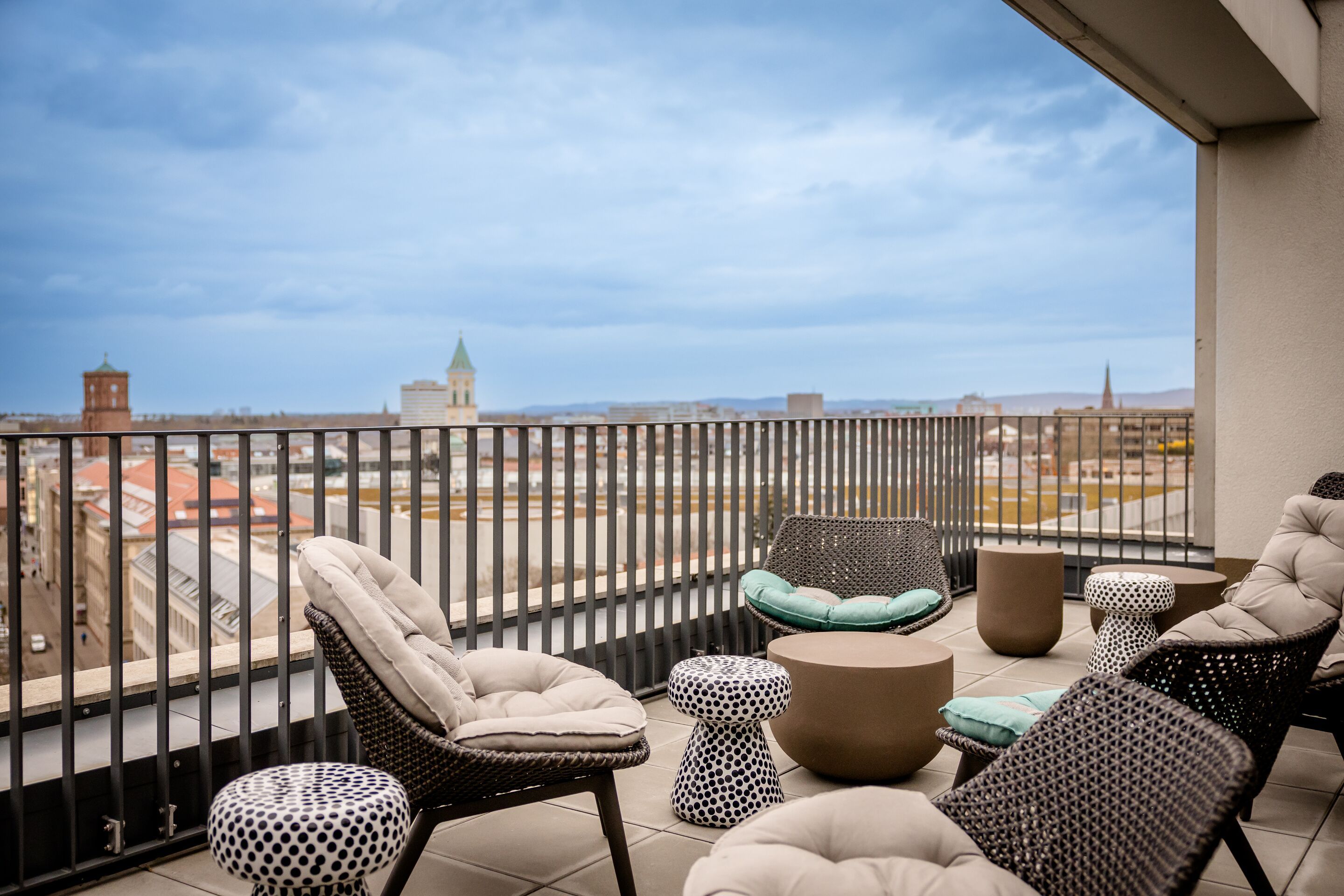 Hotel Karlsruhe Motel One Rooftop Bar Terrasse mit bequemen Polstersesseln und Tischen aus Rattan, Blick auf die Stadt und den Himmel. 
