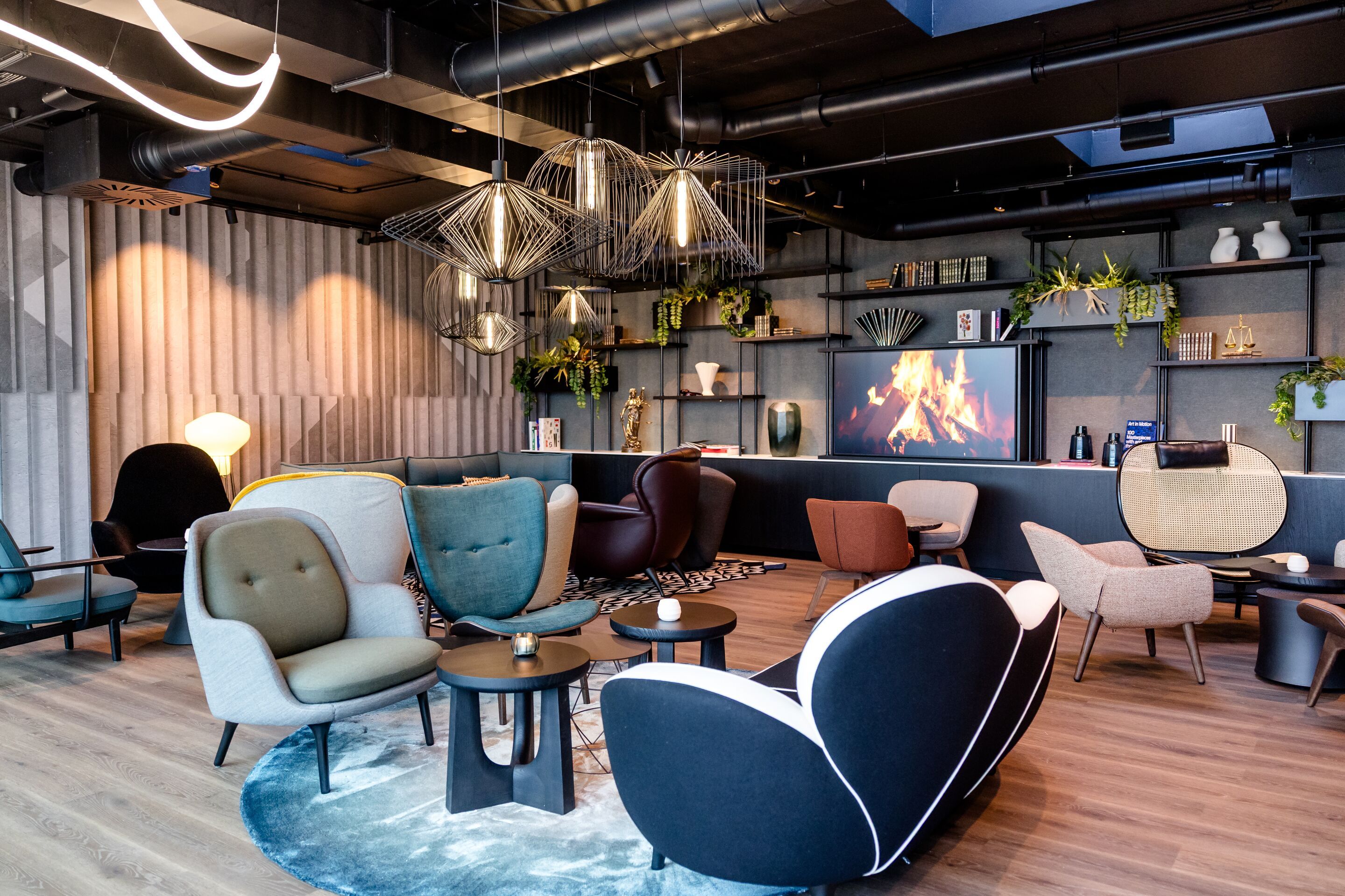 Hotel Karlsruhe Motel One Rooftop Bar Lounge mit Sesseln, Tischen und einer gemütlichen Atmosphäre, akzentuiert durch ein digitales Kaminfeuer und dekorative Pflanzen.