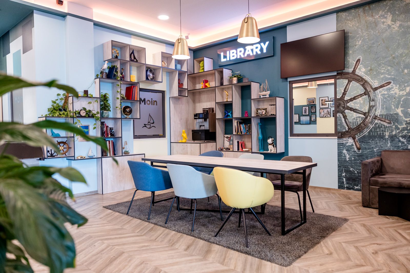 Modern eingerichteter Lese- und Arbeitsbereich mit bunten Stühlen, großem Tisch, Regalen voller Bücher und Dekoration sowie maritimem Design. Helle Atmosphäre, ideal zum Entspannen oder Arbeiten.