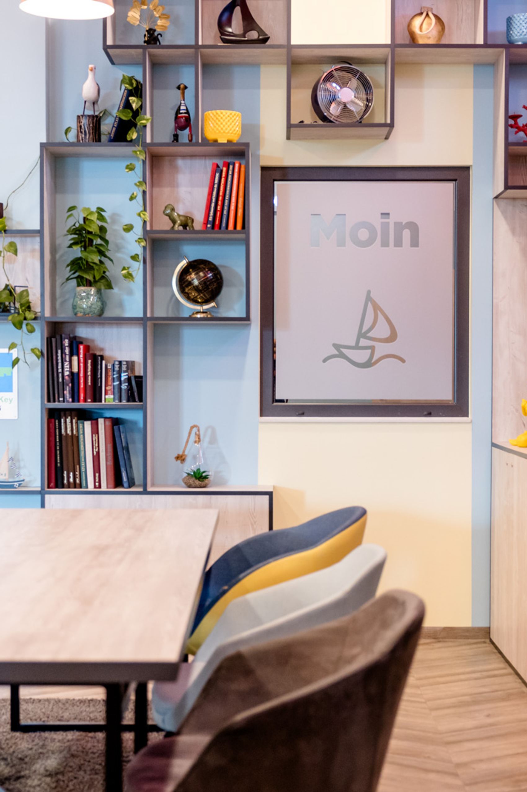 Moderner Gemeinschaftsraum mit großem Holztisch, bunten Stühlen und wandfüllendem Regal mit Dekorationen, Büchern und Pflanzen. Helle, freundliche Atmosphäre lädt zum Verweilen und Arbeiten ein.