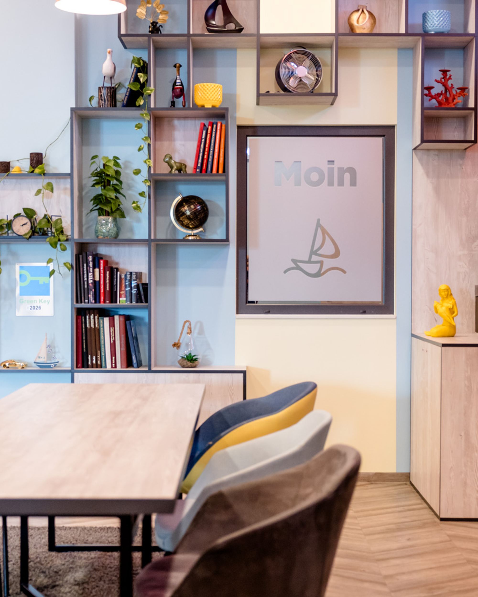 Moderner Gemeinschaftsraum mit großem Holztisch, bunten Stühlen und wandfüllendem Regal mit Dekorationen, Büchern und Pflanzen. Helle, freundliche Atmosphäre lädt zum Verweilen und Arbeiten ein.