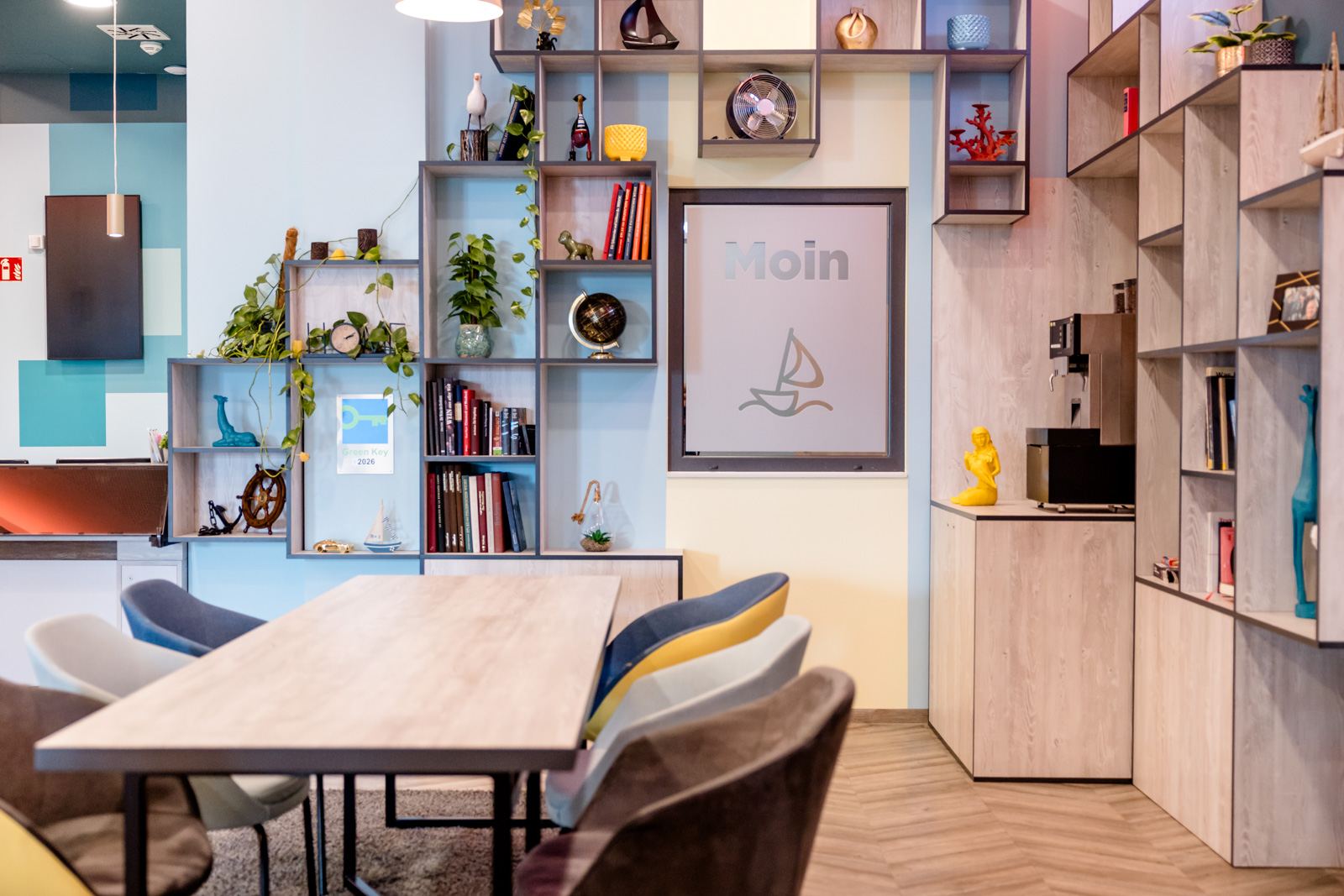 Moderner Gemeinschaftsraum mit großem Holztisch, bunten Stühlen und wandfüllendem Regal mit Dekorationen, Büchern und Pflanzen. Helle, freundliche Atmosphäre lädt zum Verweilen und Arbeiten ein. Moderner Gemeinschaftsraum mit großem Holztisch, bunten Stühlen und wandfüllendem Regal mit Dekorationen, Büchern und Pflanzen. Helle, freundliche Atmosphäre lädt zum Verweilen und Arbeiten ein.