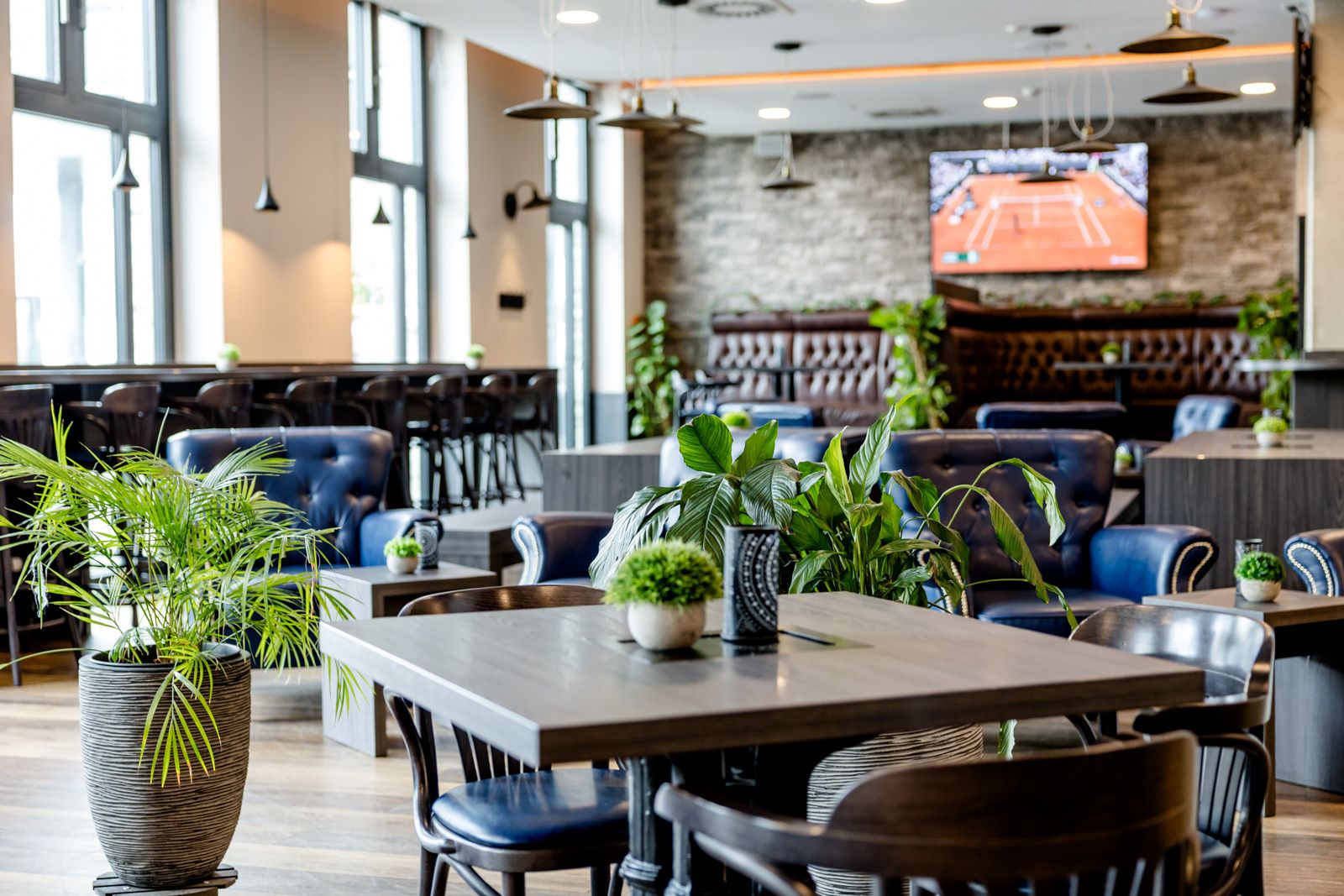 Modern eingerichteter Hotel-Lounge-Bereich mit bequemen Sesseln, Tischen und viel Tageslicht. Pflanzen sorgen für eine angenehme Atmosphäre. Im Hintergrund läuft ein Tennisspiel auf einem Fernseher, ideal zum Entspannen und Verweilen.