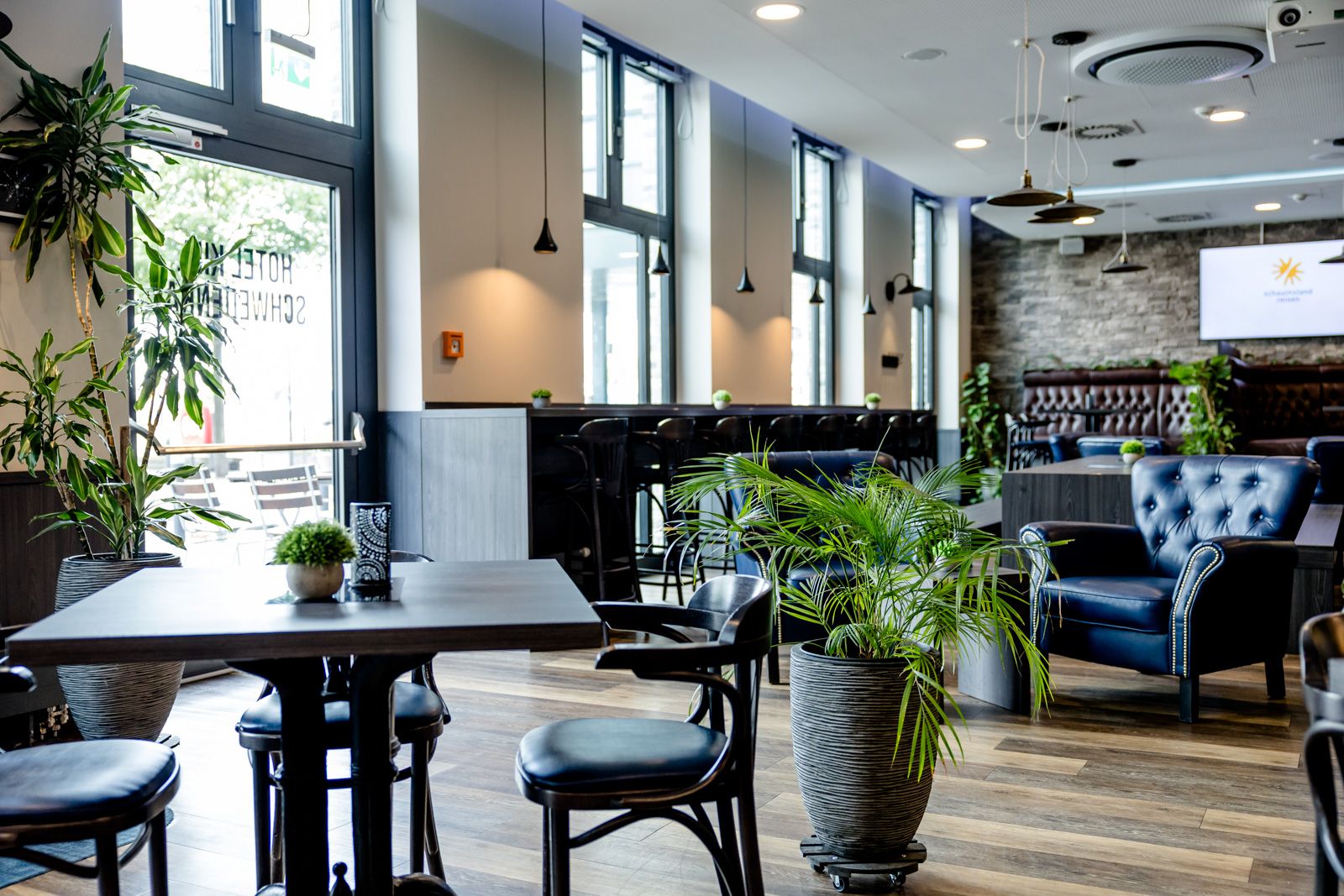 Helle, moderne Lounge mit großen Fenstern, bequemen Ledersesseln und stilvollen Holztischen. Grüne Pflanzen sorgen für eine entspannte Atmosphäre, ideal zum Entspannen oder für informelle Treffen in angenehmem Ambiente.
