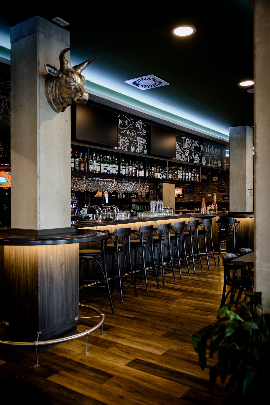 Moderne Hotelbar mit langer Holztheke, zahlreichen Barhockern und gut bestücktem Regal mit diversen Spirituosen. Warme Beleuchtung und gemütliche Atmosphäre für entspannte Abende.