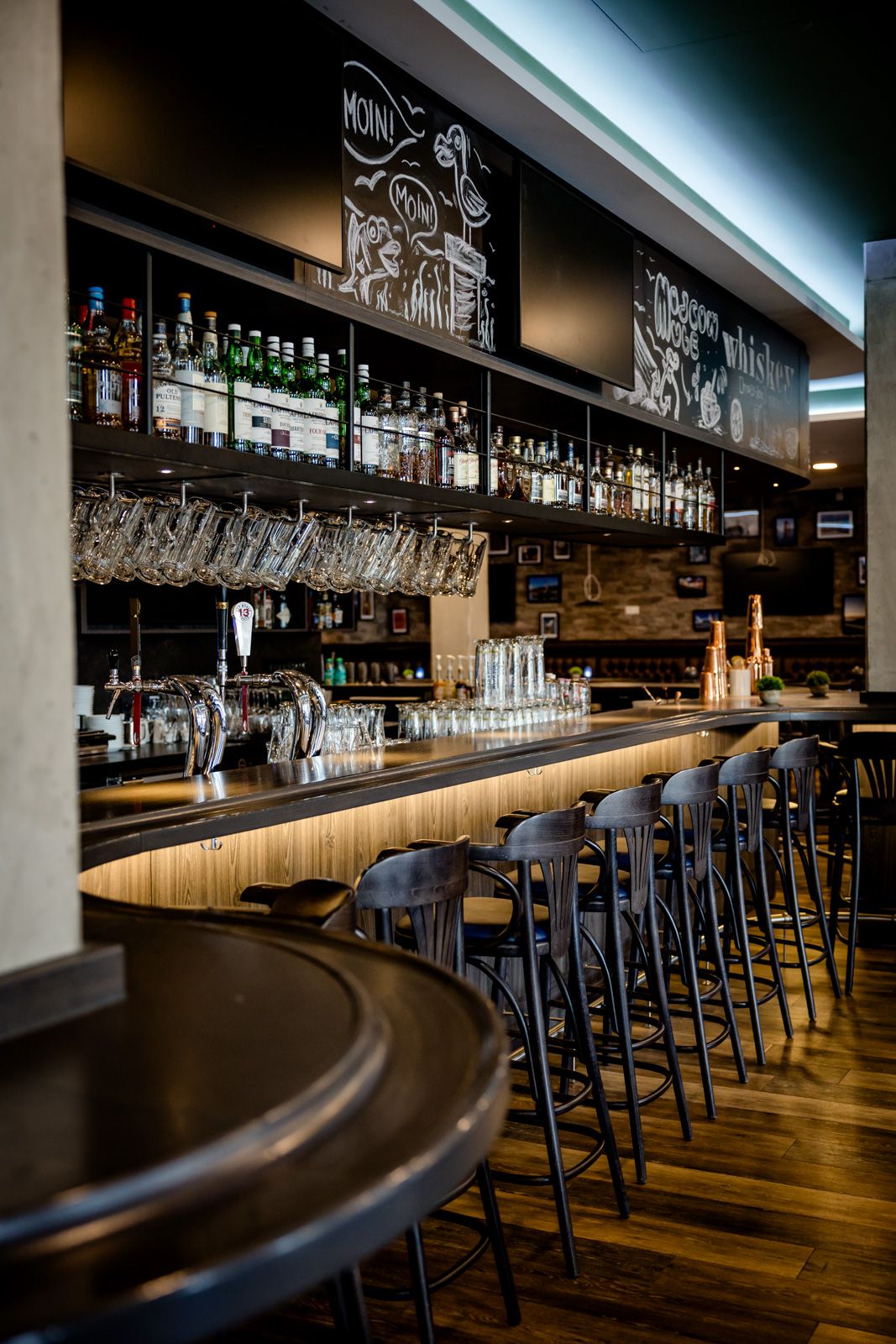 Moderne Hotelbar mit langem Holztresen, hohen Barhockern und zahlreichen Spirituosenflaschen. Gemütliche Atmosphäre mit warmem Licht und dekorativen Tafelbildern über der Bar. Ideal für entspannte Abende und gesellige Treffen.
