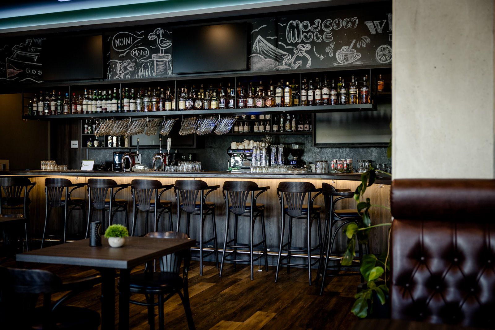 Moderne Hotelbar mit Holztresen, hohen Barhockern und einer großen Auswahl an Spirituosen. Gemütliche Atmosphäre mit dunklem Mobiliar und dekorativen Pflanzen, ideal für entspannte Abende und gesellige Treffen.