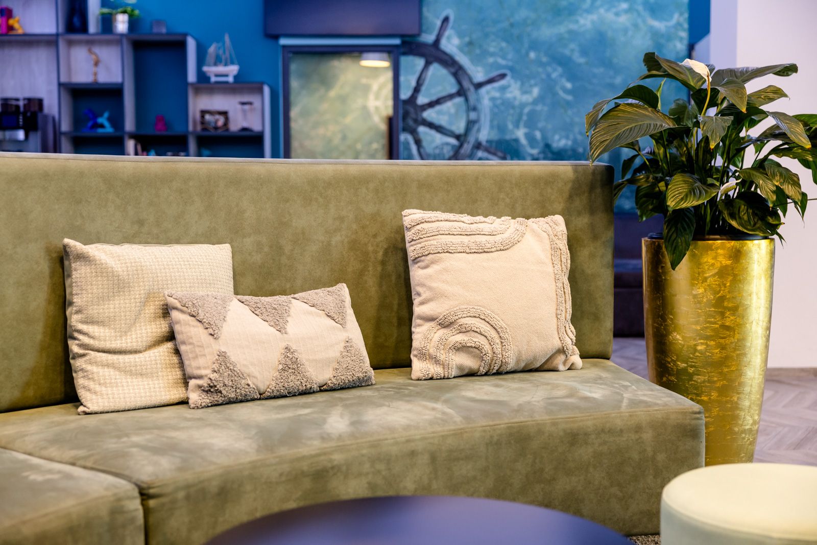 Gemütliche Sitzecke mit grünem Samtsofa und dekorativen Kissen, daneben eine hohe goldene Blumenvasen mit grüner Pflanze, im Hintergrund dezente maritime Wandgestaltung für entspannte Atmosphäre.
