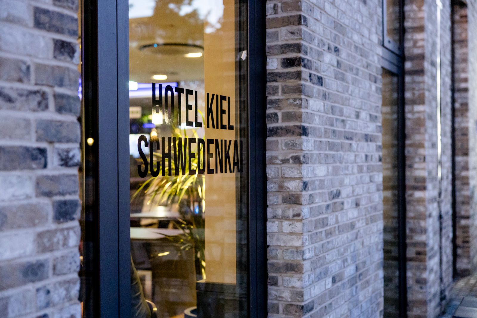 Eingangstür eines modernen Hotels mit dem Schriftzug „Hotel Kiel Schwedenkai“ auf dem Glas, umgeben von einer Fassade aus grau-beigen Ziegelsteinen. Im Inneren sind warme Lichtquellen und Pflanzen sichtbar.