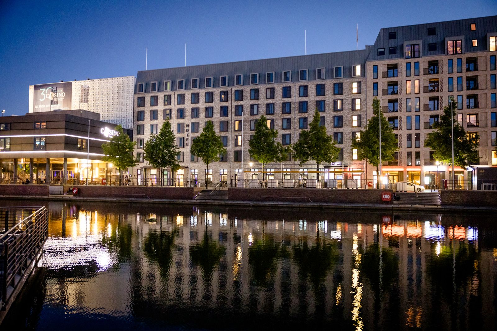 Beleuchtetes modernes Hotelgebäude am Wasser bei Abenddämmerung, mit Bäumen entlang der Promenade und ruhiger Spiegelung im Wasser.