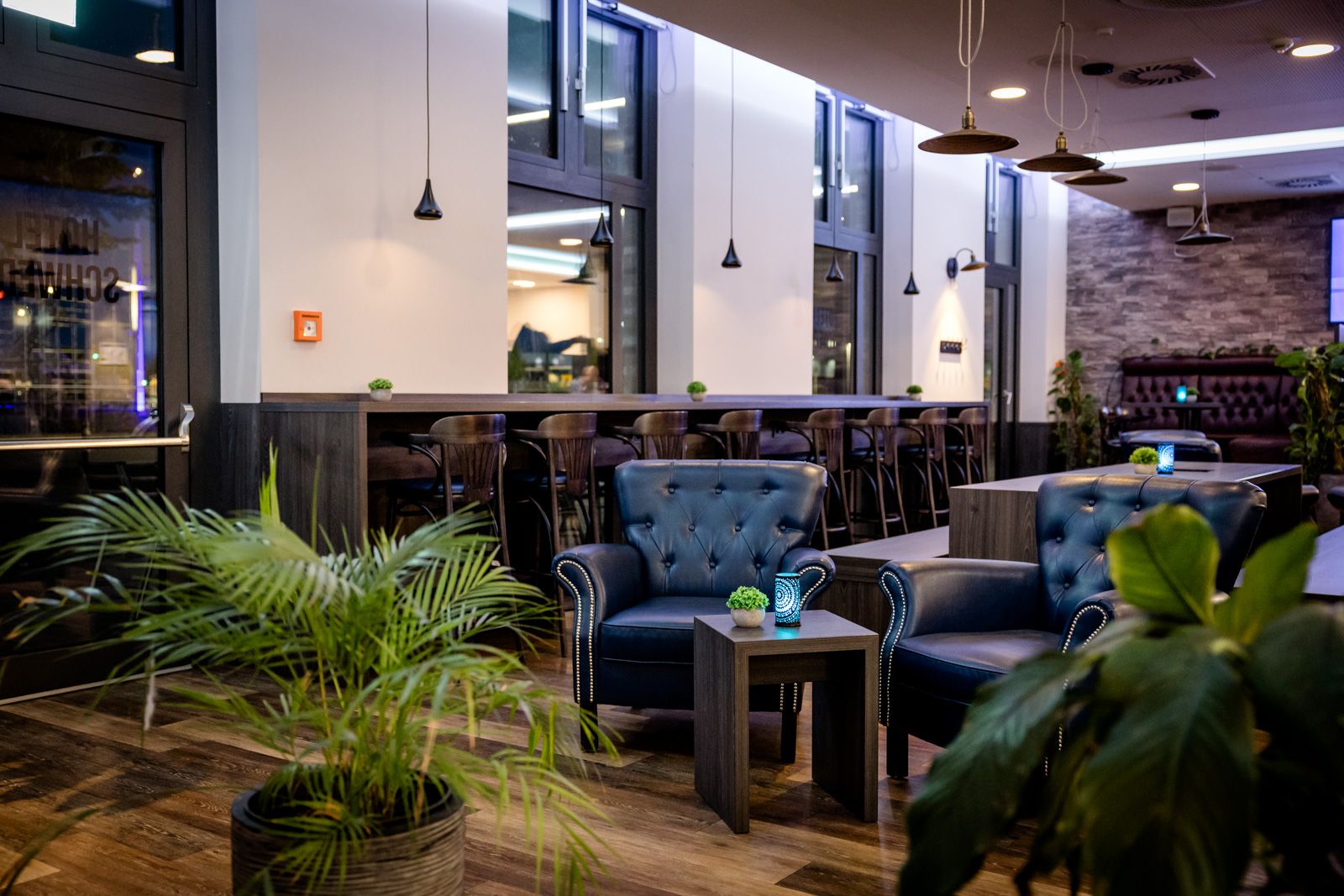 Moderne Hotel-Lounge mit bequemen Ledersesseln, kleinem Holztisch, dekorativen Pflanzen und großer Fensterfront, gemütliche Atmosphäre für entspannte Stunden oder informelle Gespräche.