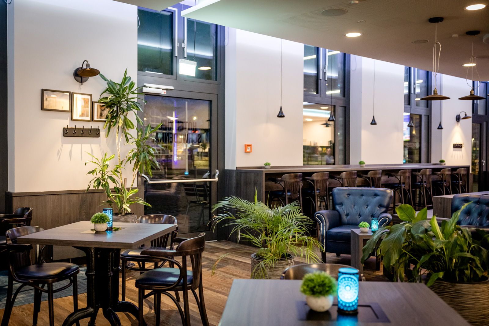 Gemütlicher Restaurantbereich mit stilvollen Holztischen, bequemen Stühlen und Sesseln, dekorativen Pflanzen und sanfter Beleuchtung, der eine entspannte Atmosphäre für Mahlzeiten und Gespräche schafft. Große Fenster lassen Tageslicht einfallen.