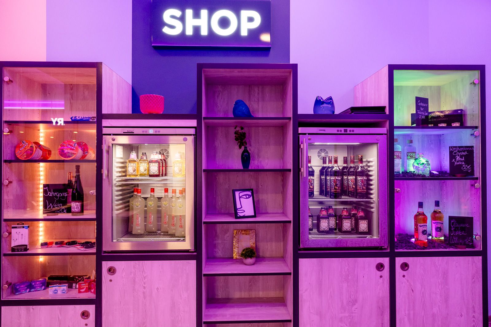 Regalbereich eines Shops mit gekühlten Getränken, Snacks und Accessoires, beleuchtet in violettem Licht, lädt zum Einkaufen ein.