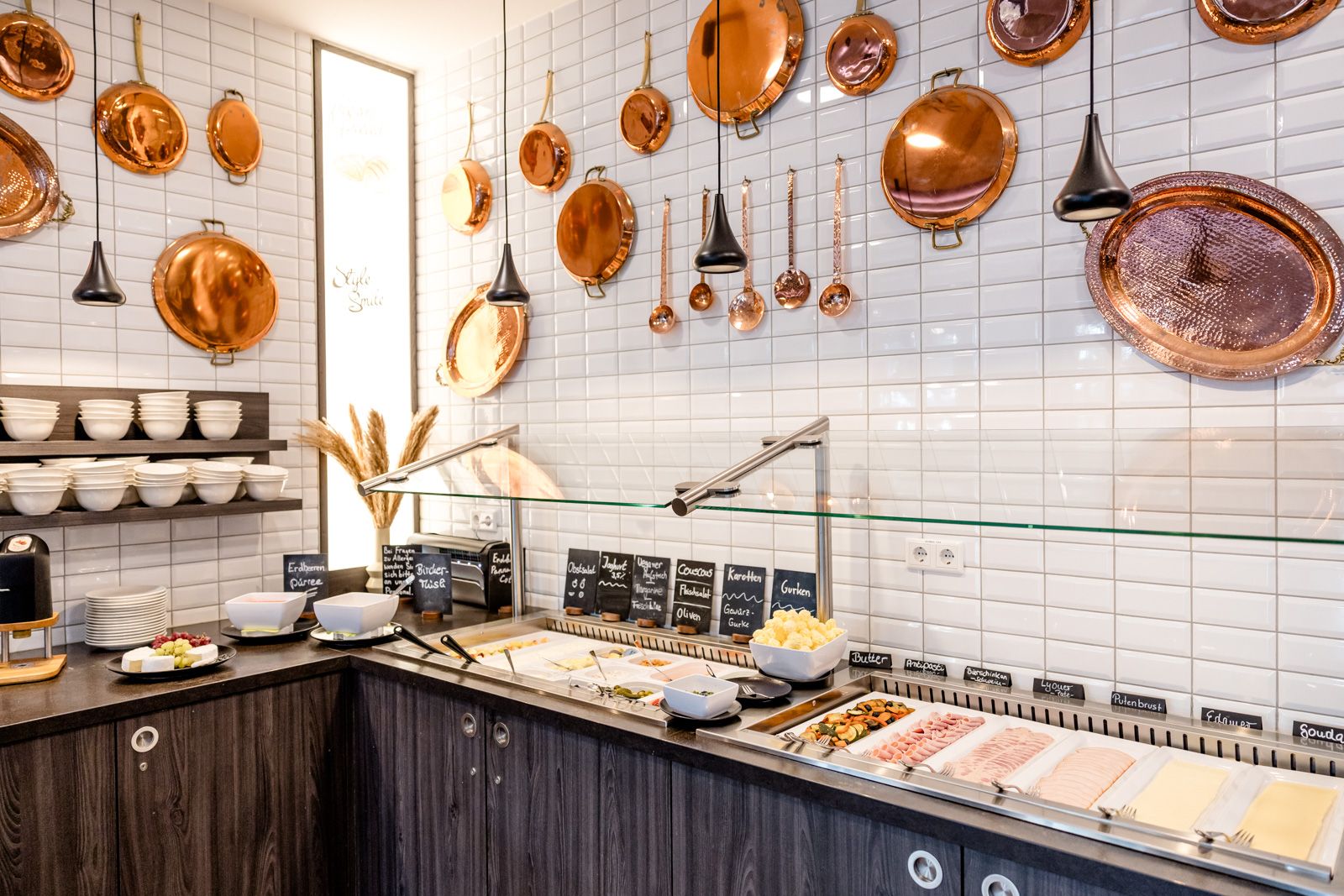 Buffetbereich mit Aufschnitt, Käse und Snacks, dekoriert mit glänzenden Kupfertöpfen an weißen Kacheln, für ein stilvolles Frühstückserlebnis im Hotel.