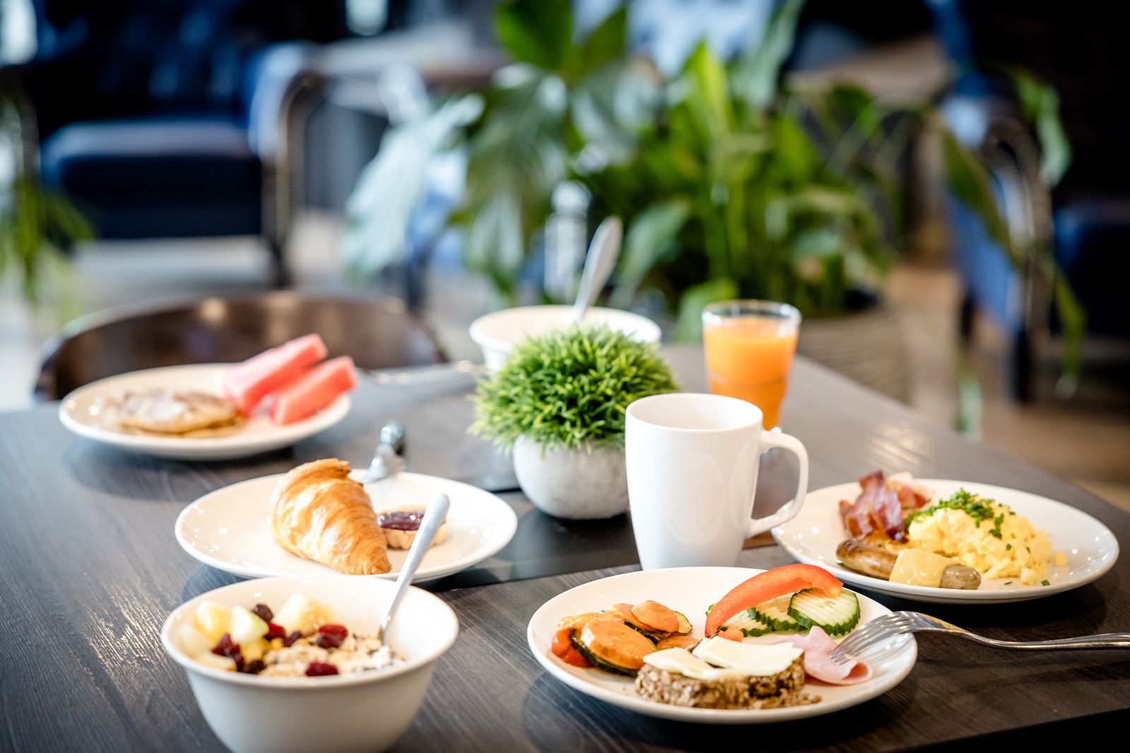Frühstückstisch mit abwechslungsreicher Auswahl: Müsli, Croissant, frisches Gemüse, Rührei mit Speck, Orangensaft und Kaffee, auf hellem Holztisch in gemütlicher Atmosphäre mit Pflanzen im Hintergrund.