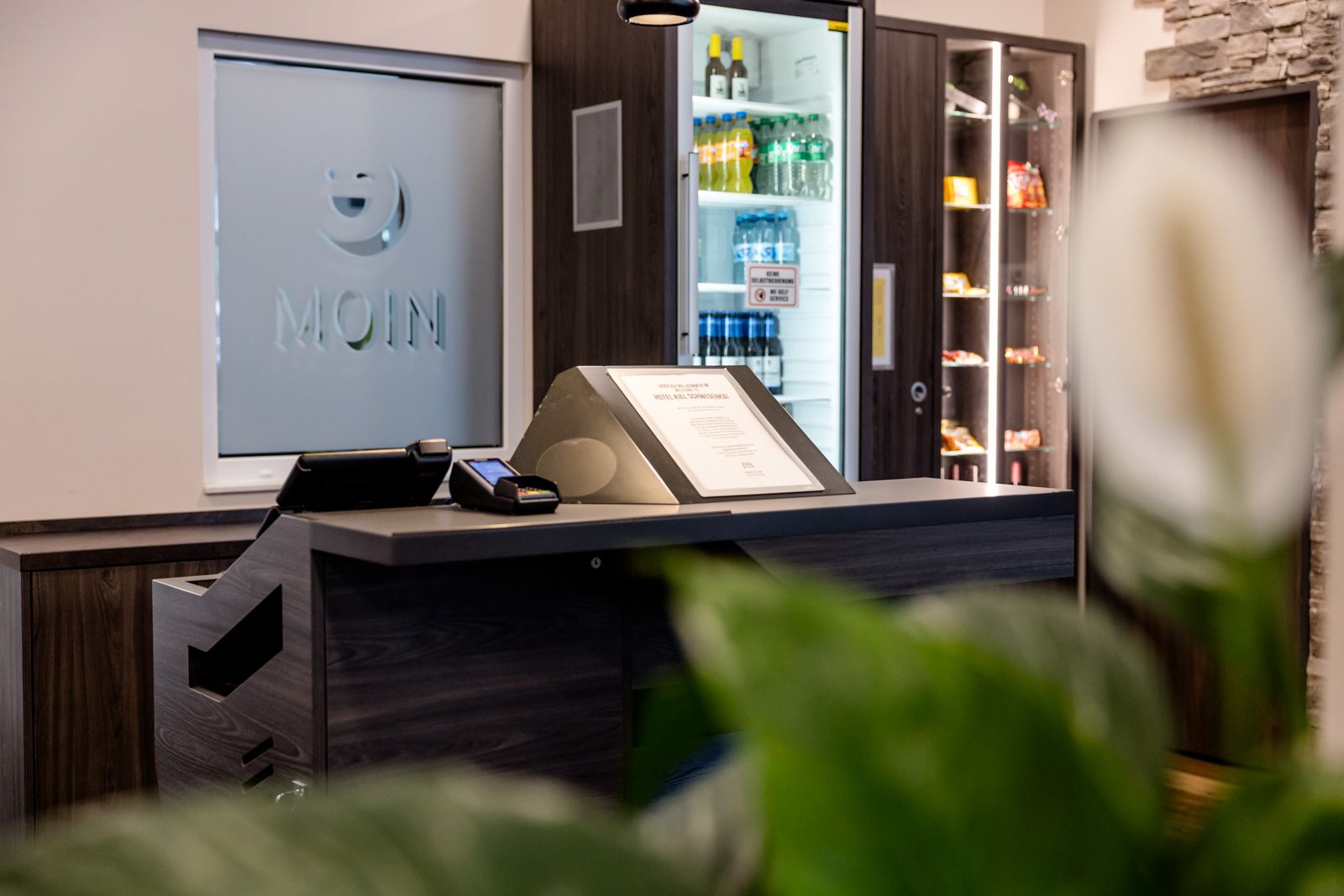 Moderne Hotelrezeption mit Kühlschrank voller Getränke und Snacks, stilvoll eingerichteter Bereich mit dunklem Holz und Steinakzenten, einladende Atmosphäre für Gästeempfang.