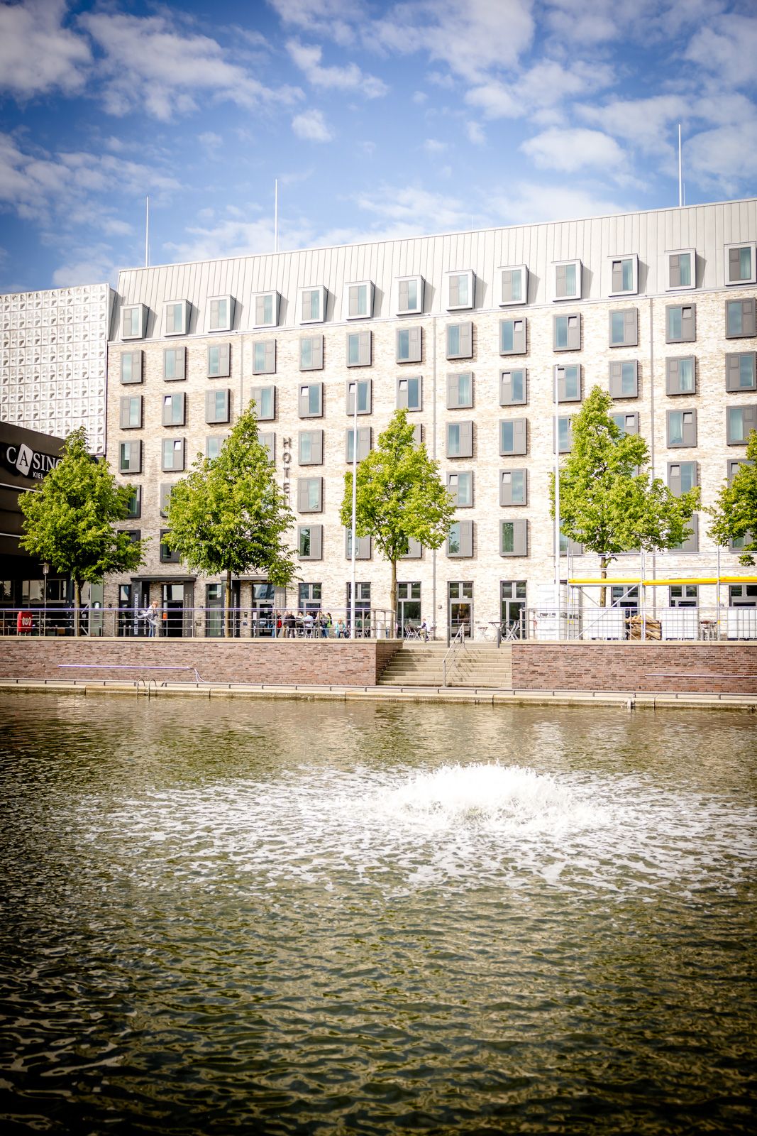 Modernes Hotelgebäude mit vielen Fenstern, davor eine Baumreihe und ein ruhiger Teich mit Wassersprudler, unter blauem Himmel mit leichten Wolken.
