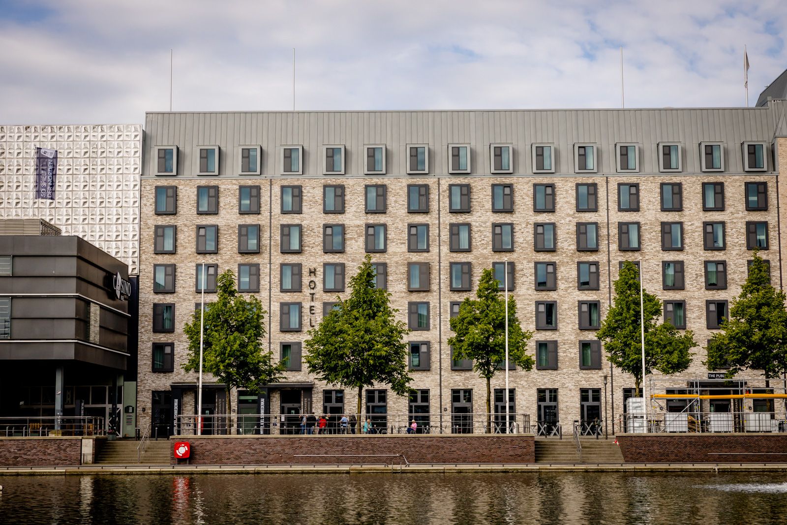 Modernes Hotelgebäude mit mehreren Stockwerken und gleichmäßig verteilten Fenstern, direkt an einem ruhigen Wasserkanal gelegen, umgeben von grünen Bäumen, ideal für entspannte Städtereisen und komfortable Unterkunft.