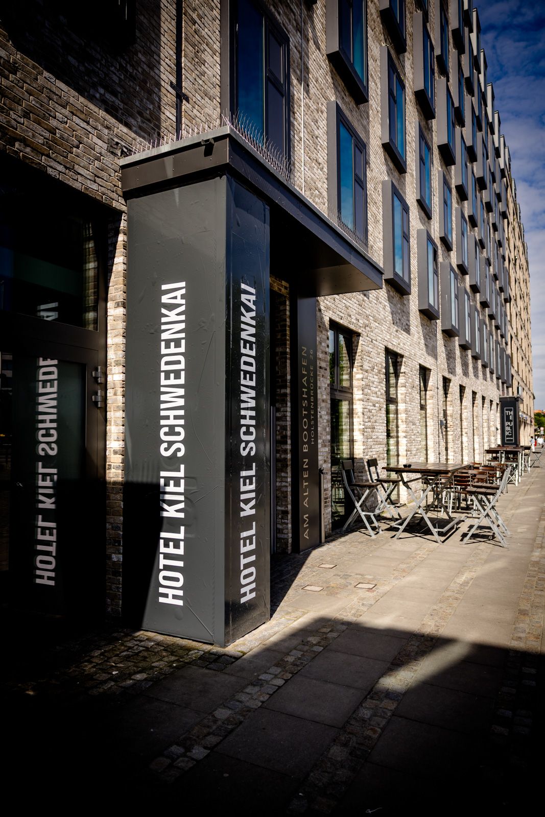 Modernes Hotelgebäude mit heller Backsteinfassade, großen Fenstern und einem Eingangsbereich mit dem Schriftzug „Hotel Kiel Schwedenkai“. Außen an der Hauswand stehen Tische und Stühle für Gäste. Sonniger Tag mit blauem Himmel.