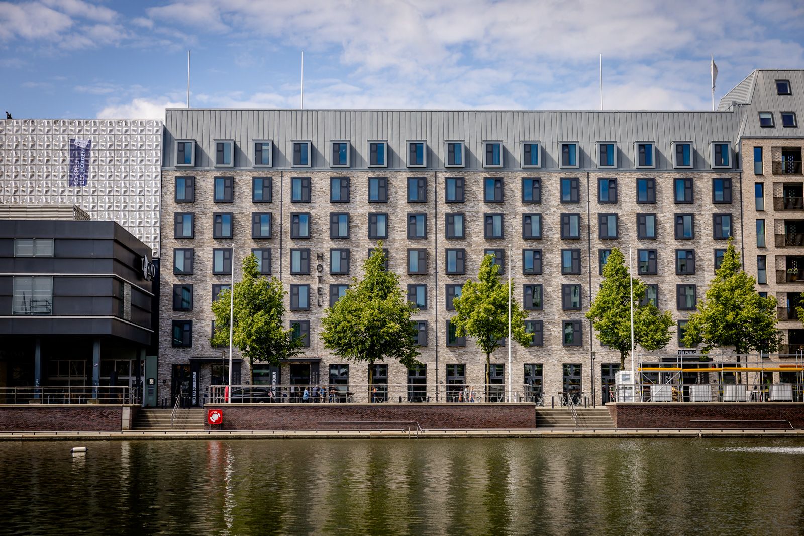 Moderner Hotelbau direkt an einem ruhigen Flussufer mit gleichmäßig angeordneten Fenstern und einer Baumreihe entlang der Promenade, die eine entspannte Atmosphäre für Geschäfts- und Freizeitgäste schafft.