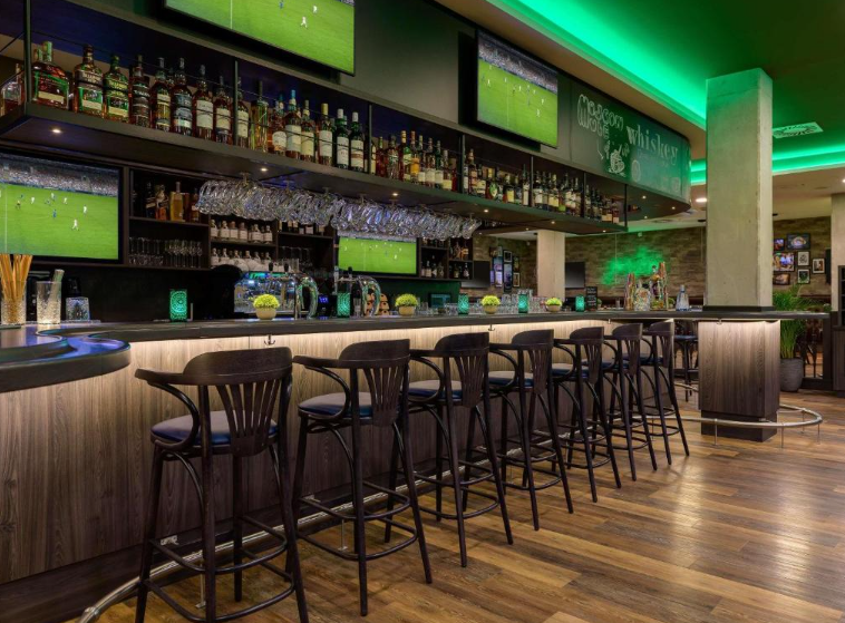 Moderne Bar mit Holzboden, beleuchtetem Tresen und mehreren Bildschirmen, die Sportübertragungen zeigen. Komfortable Barhocker, gut bestückte Regale mit Spirituosen und Glasaufhängungen für ein gemütliches, entspanntes Ambiente.