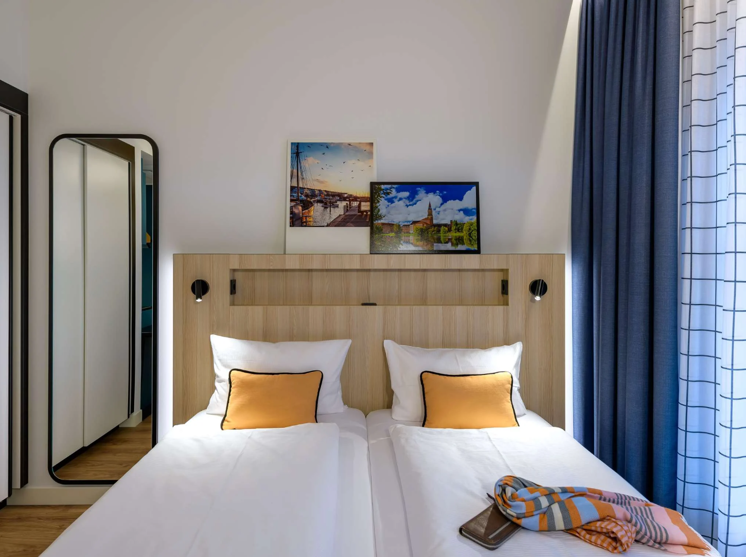 Helles, modernes Doppelzimmer mit zwei Einzelbetten, weißen Bettwaren und orangenen Kissen, dekoriert mit Stimmungsbildern an der Wand. Große Fenster mit Vorhängen lassen Tageslicht herein, neben einem schmalen Ganzkörperspiegel, das Zimmer wirkt einladend und komfortabel.