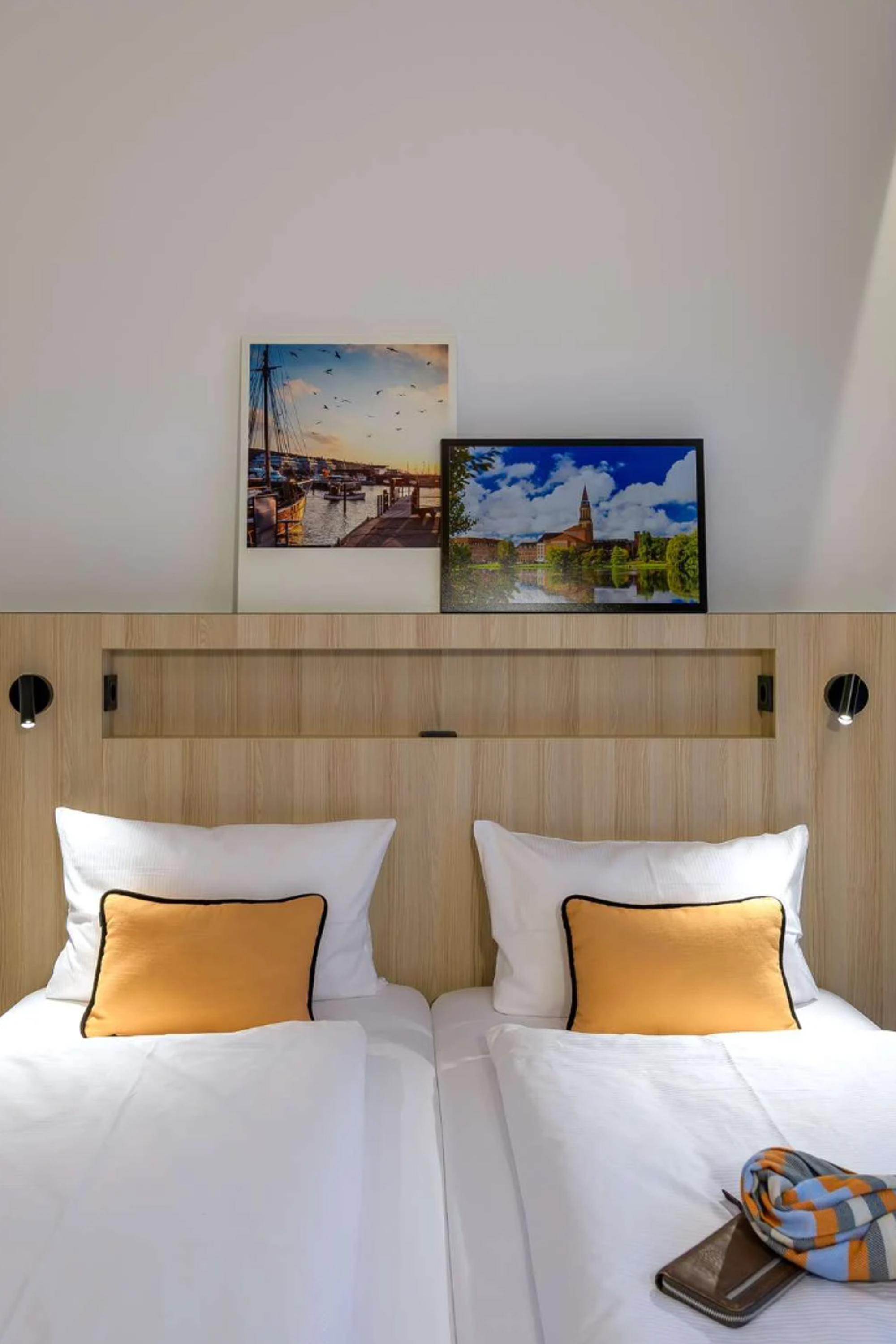 Helles, modernes Doppelzimmer mit zwei Einzelbetten, weißen Bettwaren und orangenen Kissen, dekoriert mit Stimmungsbildern an der Wand. Große Fenster mit Vorhängen lassen Tageslicht herein, neben einem schmalen Ganzkörperspiegel, das Zimmer wirkt einladend und komfortabel.