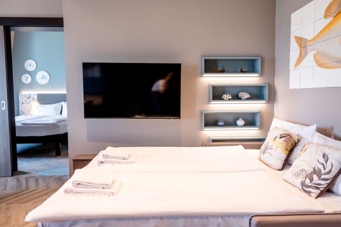 Modern eingerichtetes Hotelzimmer mit großem Doppelbett, weichen Kissen und sauberer, heller Atmosphäre, TV an der Wand und dekorativen Wandregalen mit Meeresmotiven, angrenzendes Schlafzimmer sichtbar.