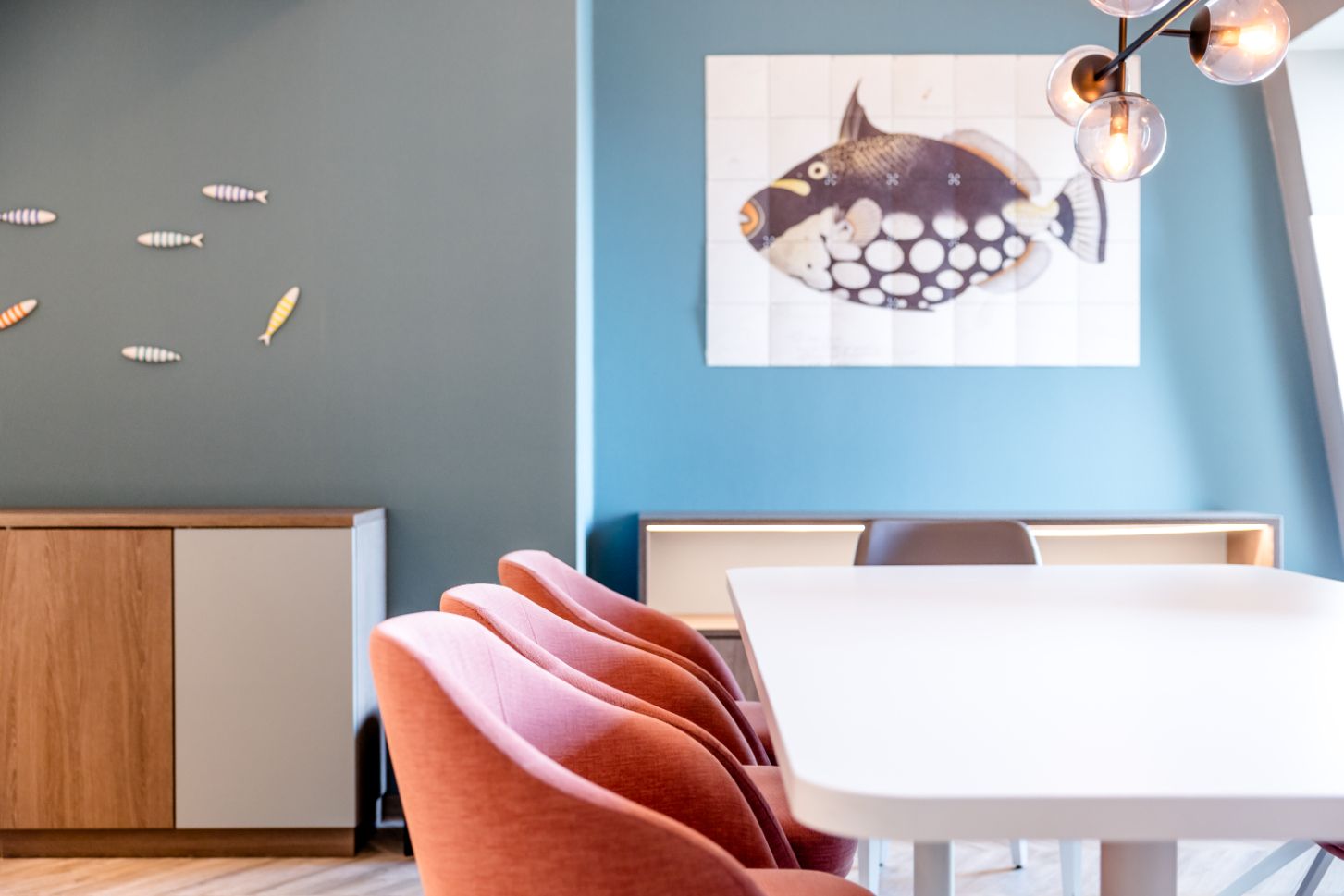 Eleganter Essbereich mit modernem weißen Tisch, stilvollen roten Stühlen und maritimen Wanddekorationen, darunter ein großes Fischbild und kleine Fische aus Metall, die eine entspannte, maritime Atmosphäre schaffen.