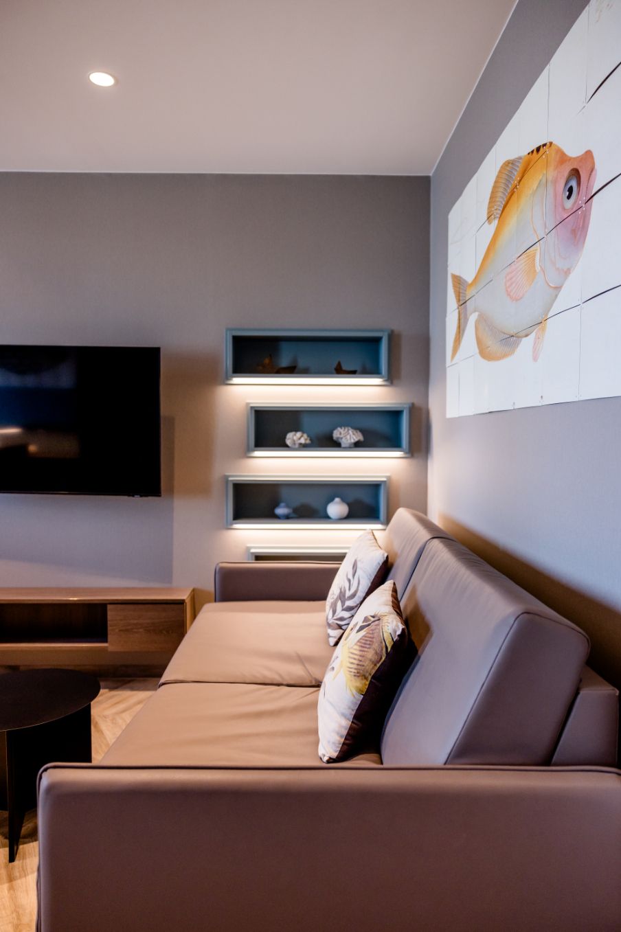 Modern eingerichtetes Wohnzimmer mit braunem Sofa, dekorativen Kissen, Wandregalen mit Muscheln und einem großen Fischbild an der grauen Wand. Gemütliche Atmosphäre für Entspannung.