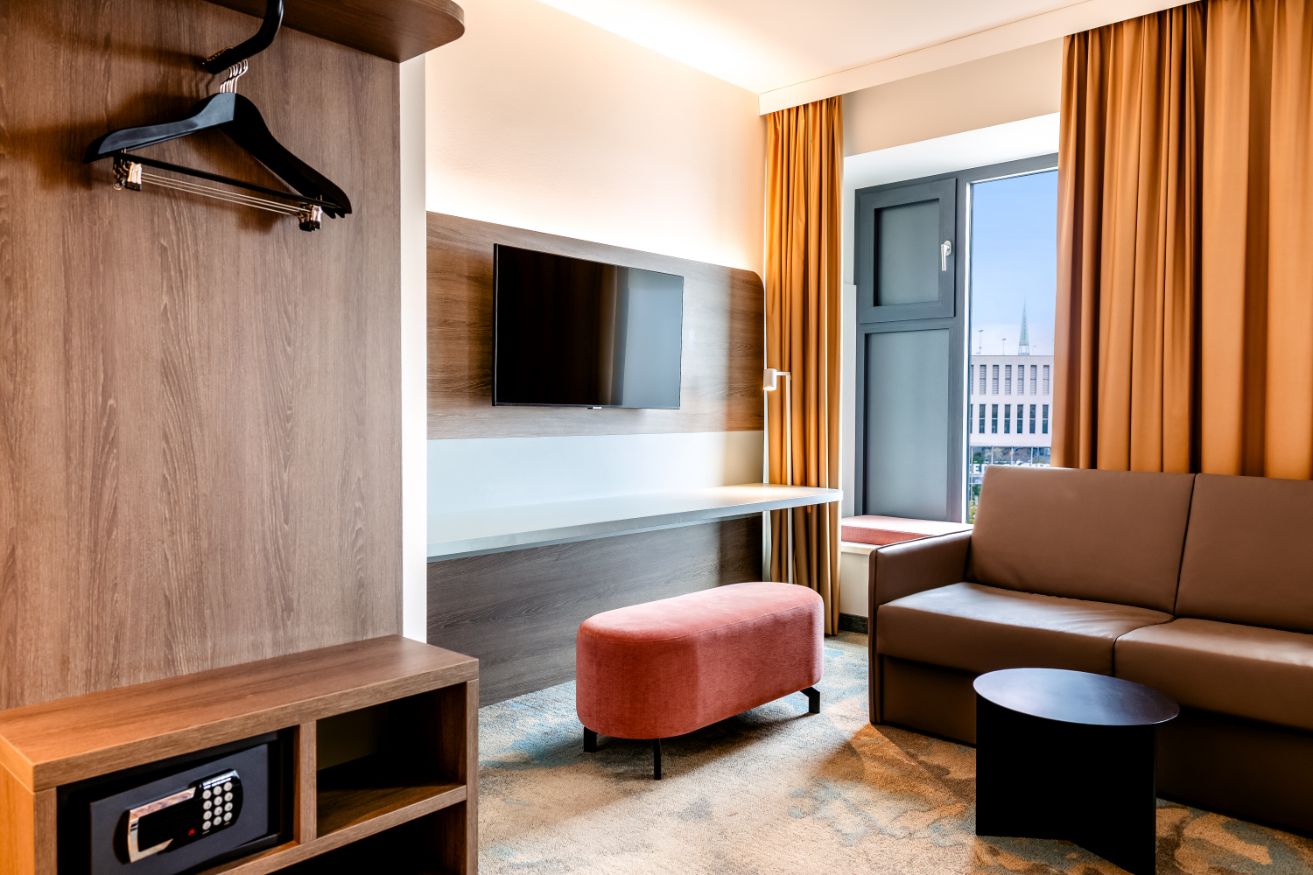 Modernes Hotelzimmer mit Sofa, Fernseher an der Wand, Fenster mit Blick auf die Stadt, Garderobe mit Kleiderbügel und Safe, helle sowie warme Farbtöne und gemütliche Atmosphäre.