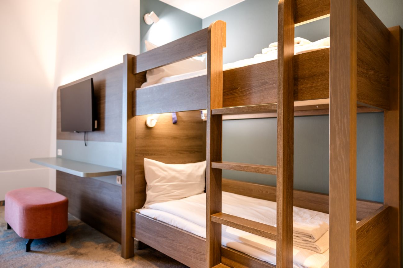 Helles Zimmer mit elegantem Holz-Etagenbett, komfortablen weißen Bettwaren, Wandlampe und moderner Wandhalterung für einen Fernseher, ideal für komfortables Übernachten in praktischem Ambiente.