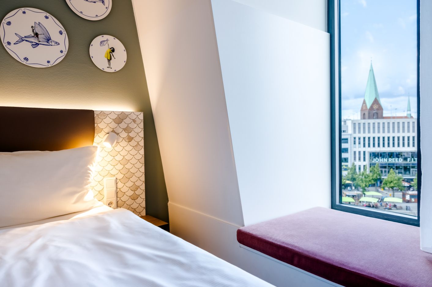 Helles, modern eingerichtetes Hotelzimmer mit gemütlichem Bett, stilvoller Wanddekoration und großem Fensterplatz mit Blick auf eine Stadt mit historischen Gebäuden und Park.