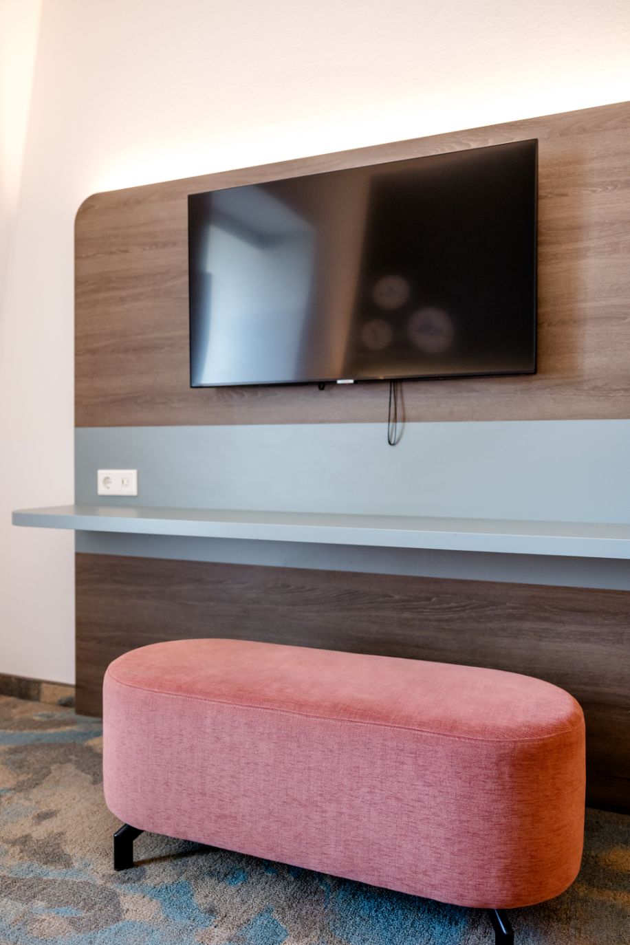 Moderner Hotelzimmerbereich mit flachem Fernseher an Holzpaneel, hellem Arbeits- oder Ablagetisch und einem gemütlichen, rosa Sitzhocker für entspannte Momente.