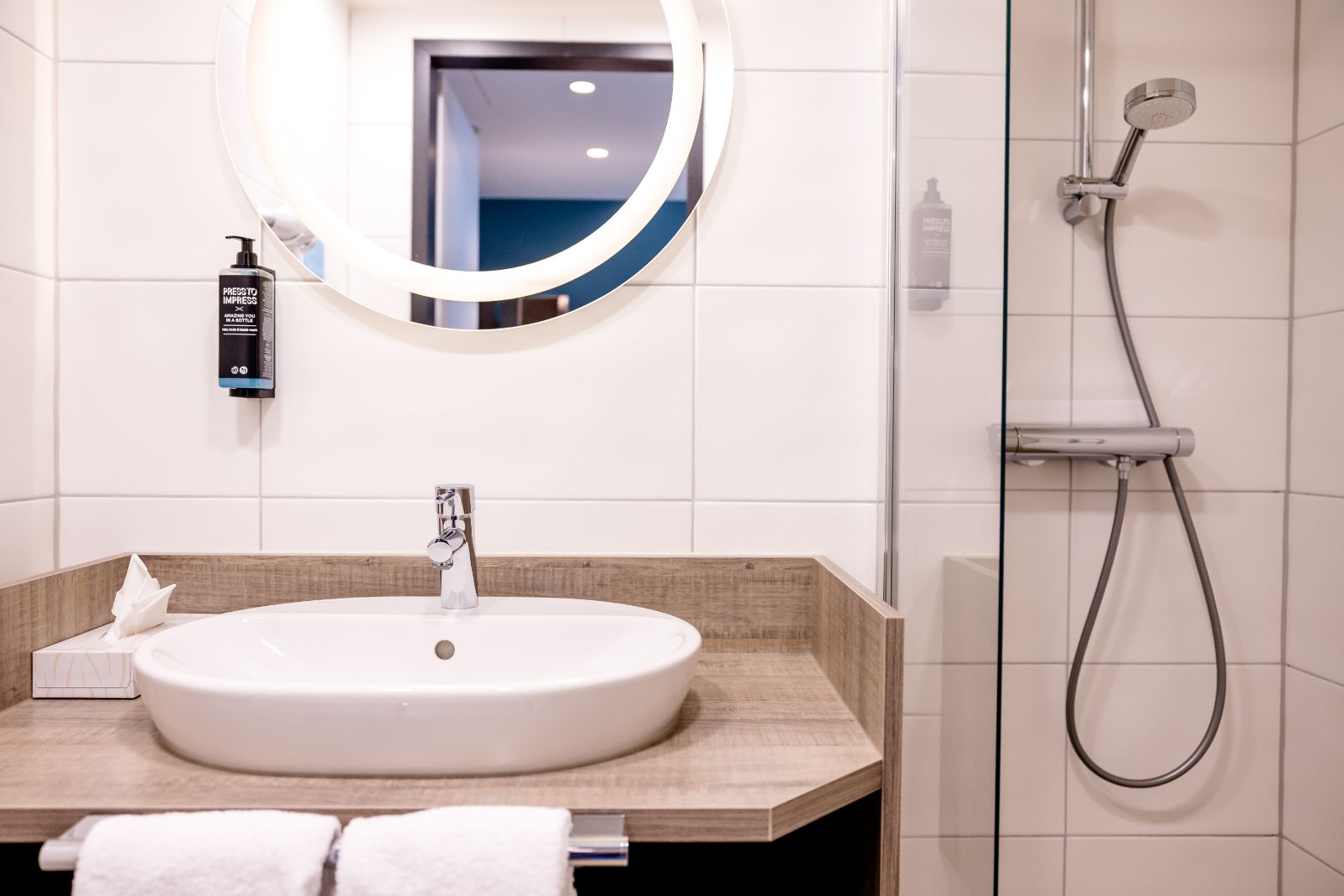 Moderne Hoteldusche mit Regenduschkopf, rundem Spiegel und weißem Waschtisch auf hellem Fliesenhintergrund, Handtücher und Seifenspender für komfortable und saubere Badnutzung.