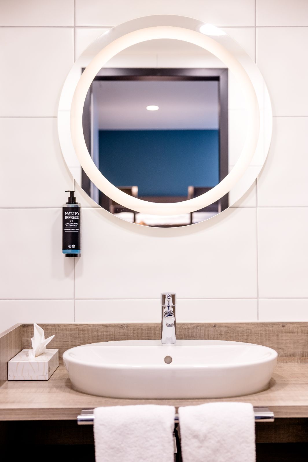 Moderner Badezimmer-Waschtisch mit rund beleuchtetem Spiegel, weißem Waschbecken, Wasserhahn und eingehängten weißen Handtüchern, flankiert von einem Taschentuchspender und einer Seifenspenderflasche.