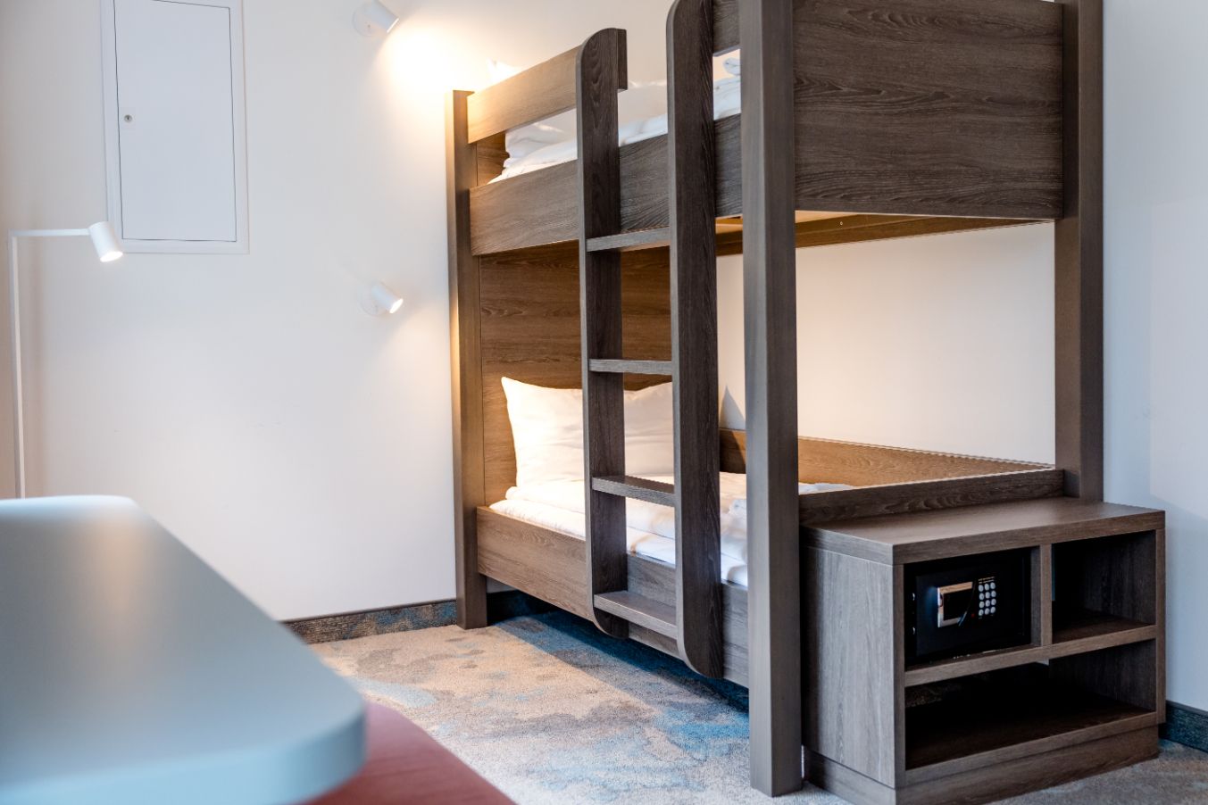 Zweistöckiges Bett aus dunklem Holz in einem hellen, modernen Zimmer mit weichem Teppichboden und integriertem Safe in einem kleinen Regal neben dem Bett.