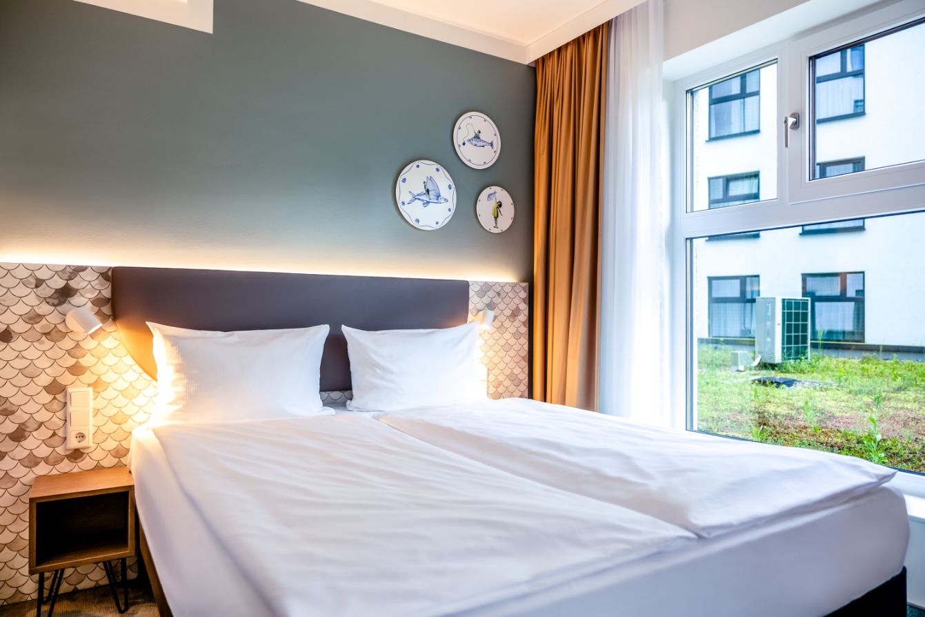 Helles, modern eingerichtetes Doppelzimmer mit großem Bett, gemütlicher Beleuchtung, dekorativen Wanduhren und großem Fenster mit Blick auf einen Innenhof.