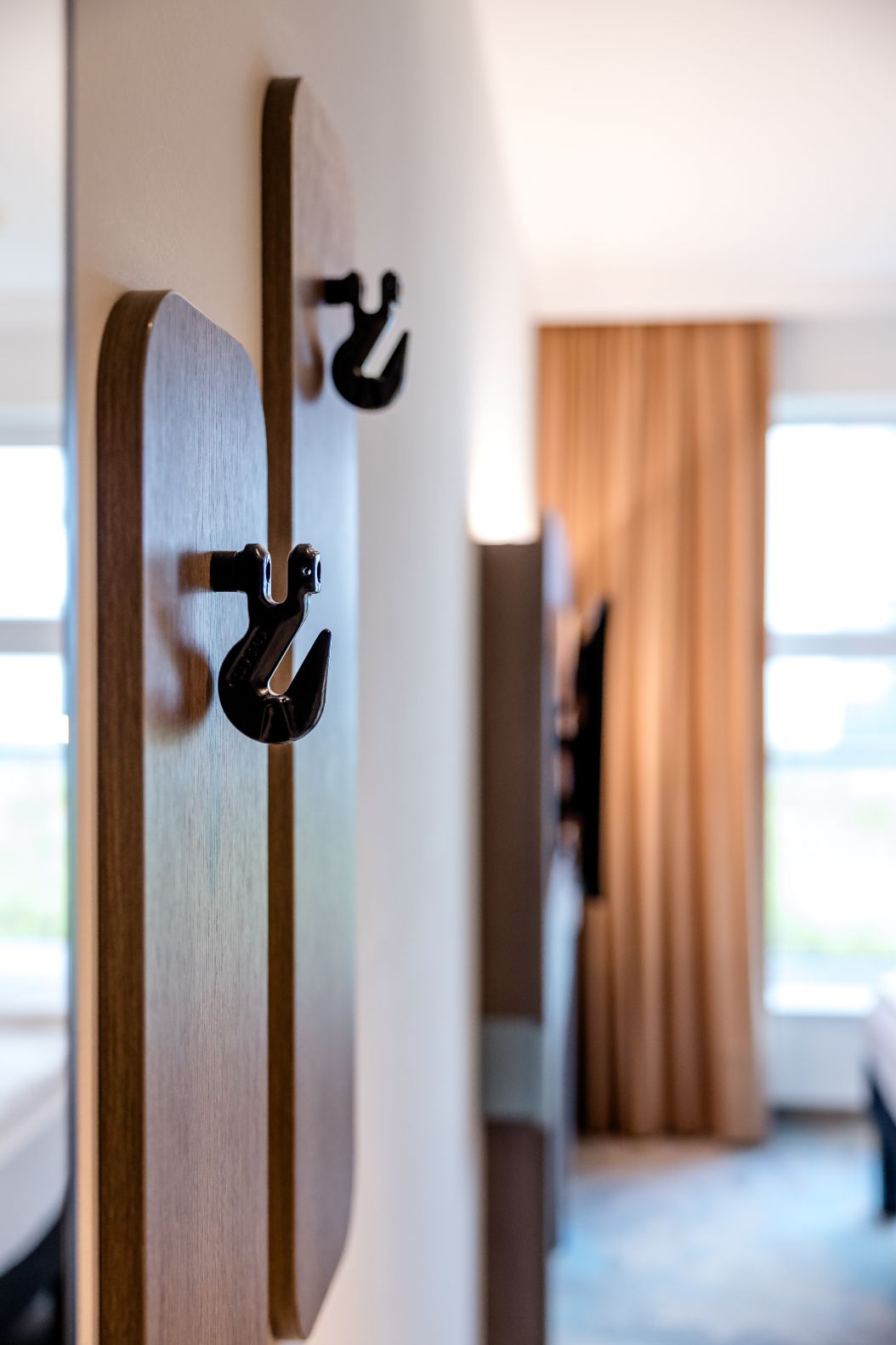 Nahaufnahme von zwei schwarzen Haken an einer Wandplatte im Hotelzimmer, im Hintergrund Tageslicht durch Fenster mit braunen Vorhängen.
