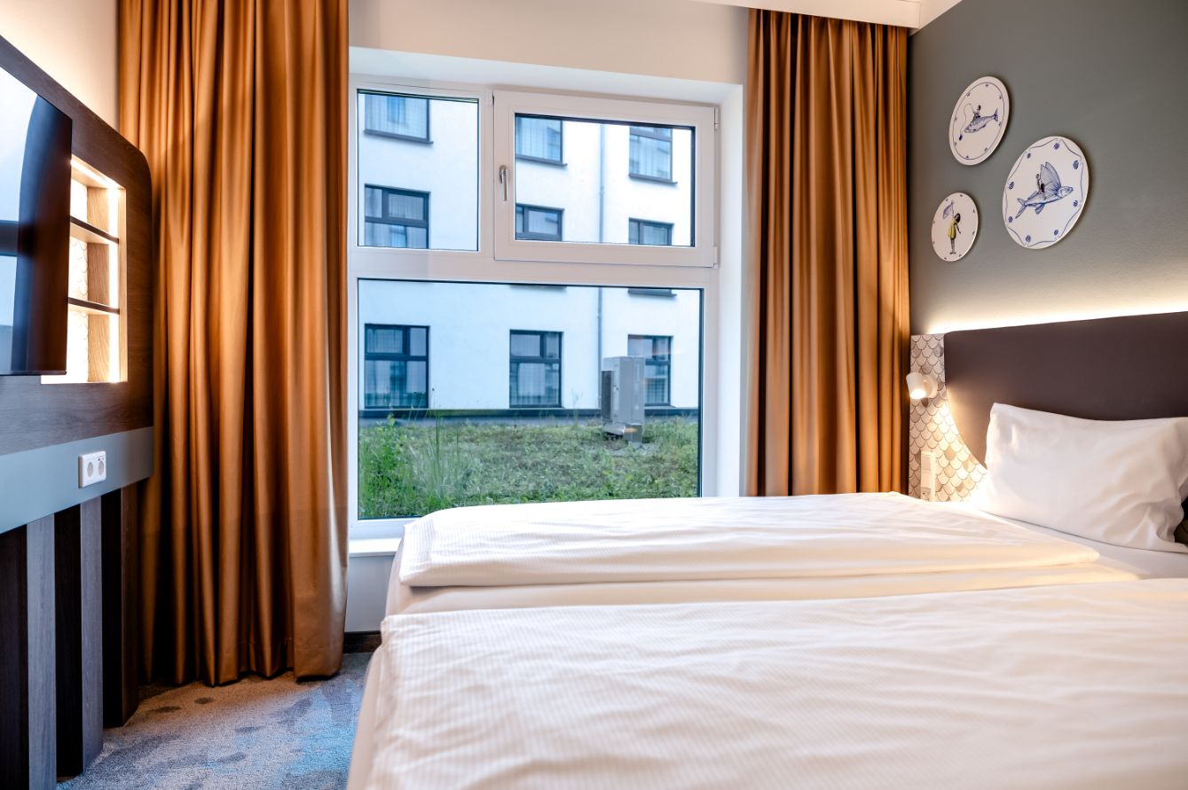 Helles Hotelzimmer mit großem Fenster und Blick auf Nachbargebäude, gemütlichem Einzelbett, warmen braunen Vorhängen und stilvoller Wanddekoration mit drei runden Bildern.