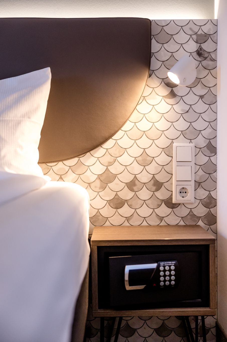 Beleuchtete Hotel-Zimmerecke mit weichem Polsterkopfteil, modernem Wandsafe, stilvoller Tapete und Leselampe für angenehme Nachtruhe und sichere Aufbewahrung persönlicher Gegenstände.