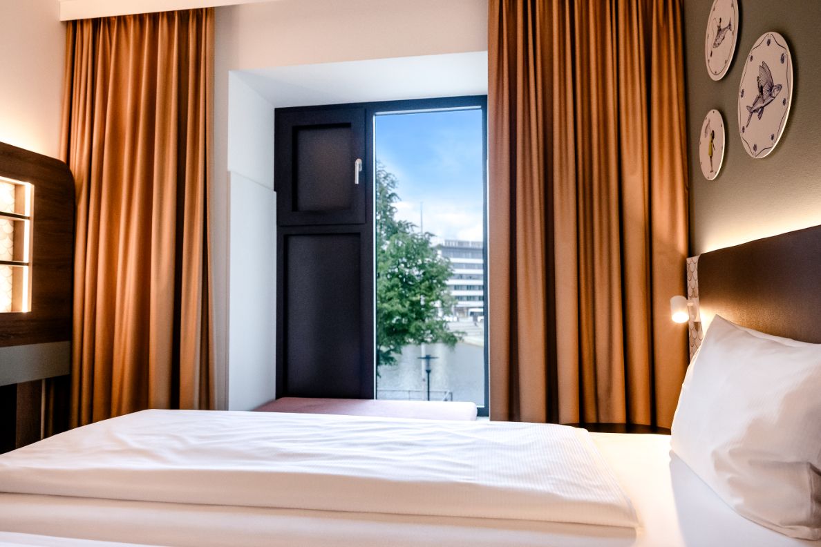 Helles Hotelzimmer mit komfortablem Bett, warmen Vorhängen und großen Fenstern, die Blick auf eine Stadtlandschaft mit Baum und Gebäuden bieten.