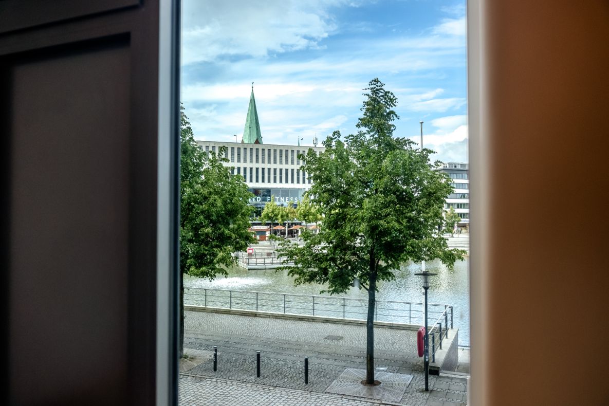 Blick aus einem Fenster auf einen ruhigen Fluss mit Uferpromenade, Bäumen und Gebäuden im Hintergrund unter blauem Himmel.