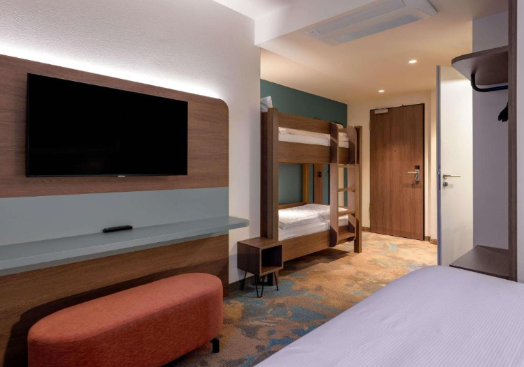 Hotel Kiel Schwedenkai Modern eingerichtetes Zimmer mit Einzelbett, Etagenbett, Fernseher und Sitzhocker, helle Beleuchtung und freundliche, warme Farben für einen komfortablen Aufenthalt.