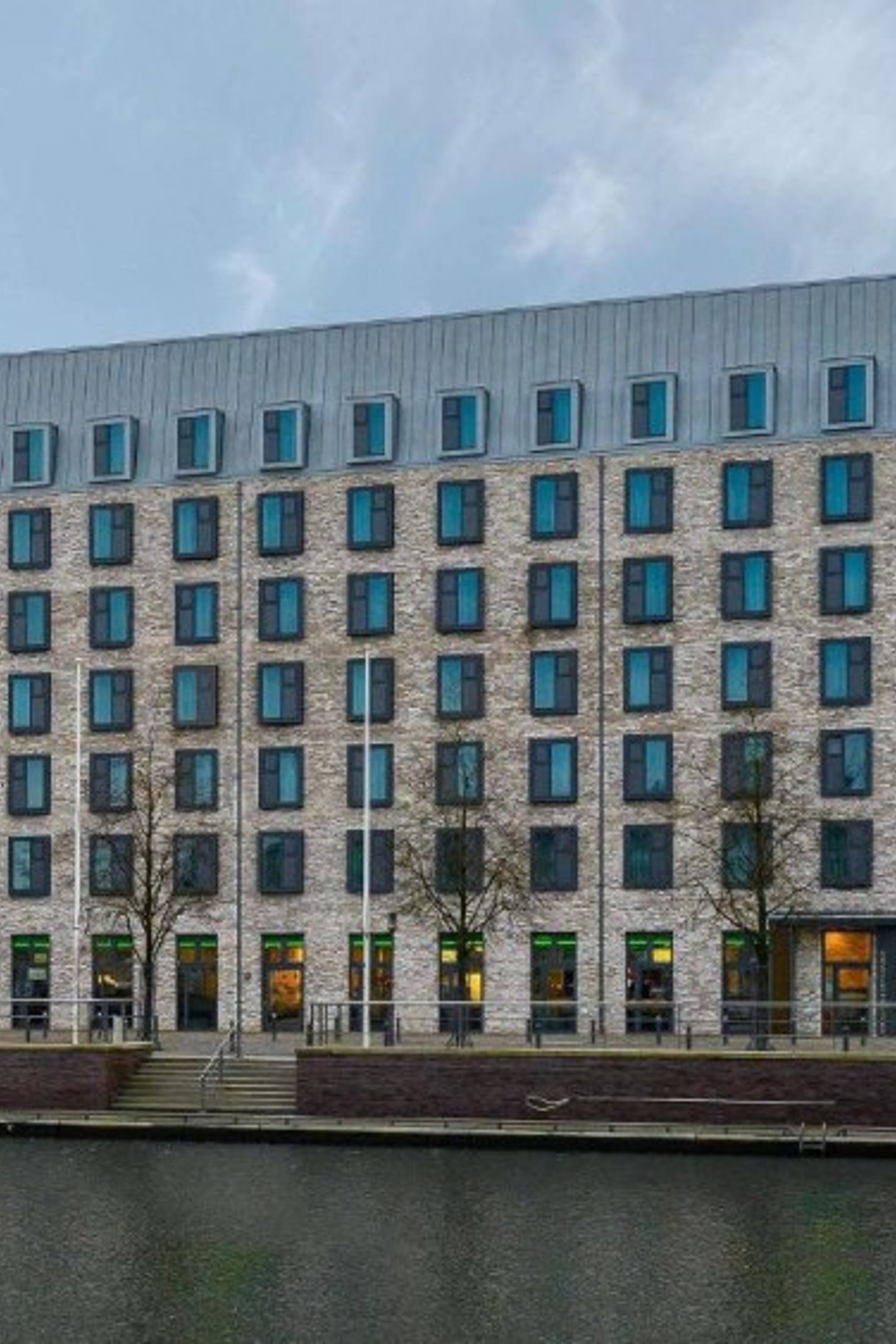 Hotel Kiel Schwedenkai Modernes Hotelgebäude mit zahlreichen Fenstern, direkt am Wasser gelegen, bei bewölktem Himmel.
