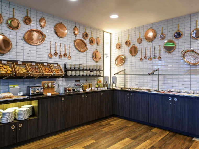 Hotel Kiel Schwedenkai Frühtücksbuffet mit Gebäck, Müsli- und Cerealienoptionen, einladendem Ambiente und dekorativen Kupfertöpfen an weiß gekachelten Wänden, für einen entspannten Start in den Tag.