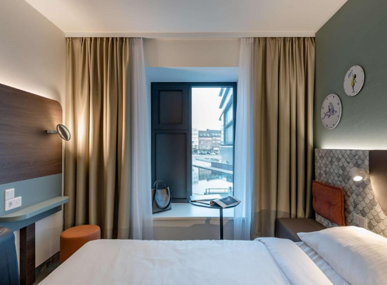Modern eingerichtetes Hotelzimmer mit gemütlichem Bett, großem Fenster mit Stadtblick, Sitzgelegenheit am Fenster und dezenten, warmen Farbtönen für eine entspannte Atmosphäre.