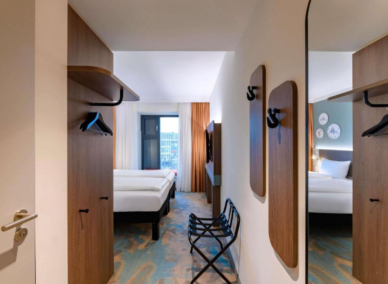 Moderner Hotelzimmer-Eingangsbereich mit offenem Kleiderschrank, Spiegeln und Blick ins helle Zimmer mit zwei Betten und großem Fenster, das Tageslicht hereinlässt. Schlichte, komfortable Einrichtung für einen angenehmen Aufenthalt.