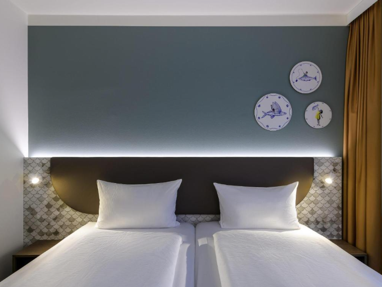 Zweibettzimmer mit klaren weißen Bettwäsche, dezent beleuchtetem Kopfteil und einer modernen Wandgestaltung mit drei dekorativen Tellermotiven über dem Bett. Gemütliche Atmosphäre für geschäftliche oder private Gäste.